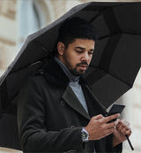 An image of a man looking at his phone in a black coat holding a black open umbrella resting on his shoulder protecting him from the rain. 