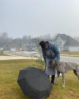 A photo of a woman petting a dog with an on a lawn in a neighborhood with an open umbrella resting on the grass. 