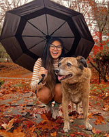 A lady crouching in the woods next to her dog holding an open umbrella that is resting on her shoulder while one hand while taking a photo with a camera with her other hand and petting her dog in the other. It is autumn and the leaves and trees are red and orange. 