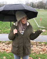 A lady standing outdoors near a soccer field holding an open umbrella that is resting on her shoulder wearing a jacket and a beanie. 