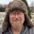 A zoomed in photo of a man standing outside in a fur hat wearing glasses. 