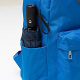 A navy compact umbrella wrapped up and tucked into the side pocked of a bright blue backpack. The umbrella's handle is exposed showing off the automatic open and close button, as well as the wrist strap.