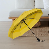 The YellowTop Best-Selling Windproof Compact Travel Umbrella is pictured open and leaning to the left. The background is an aesthetic loveseat, with a rattan pouf and a jute rug.